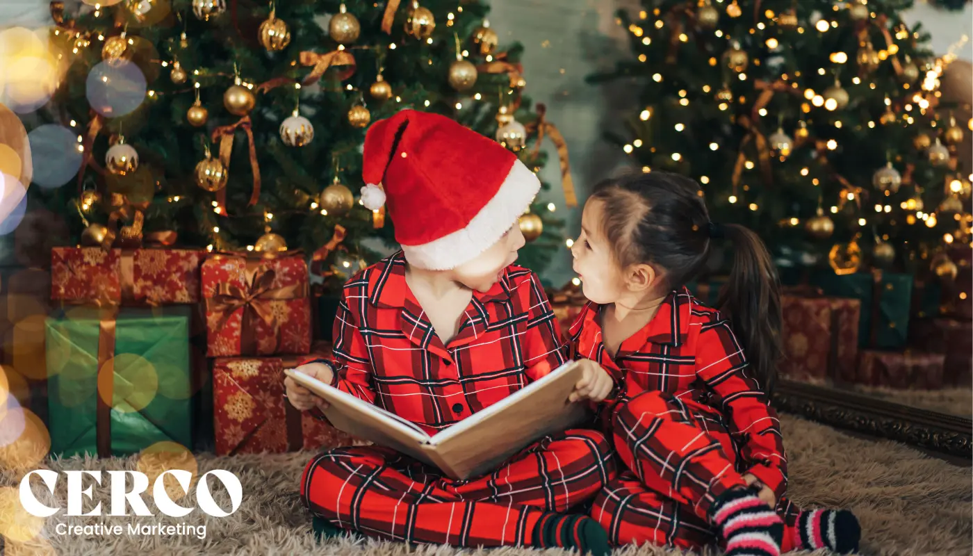 Children in Christmas pyjamas reading a book by the tree with gifts.