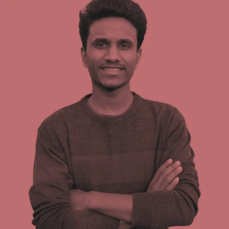 Young South Asian man wearing a striped sweater with arms crossed smiles at the camera.