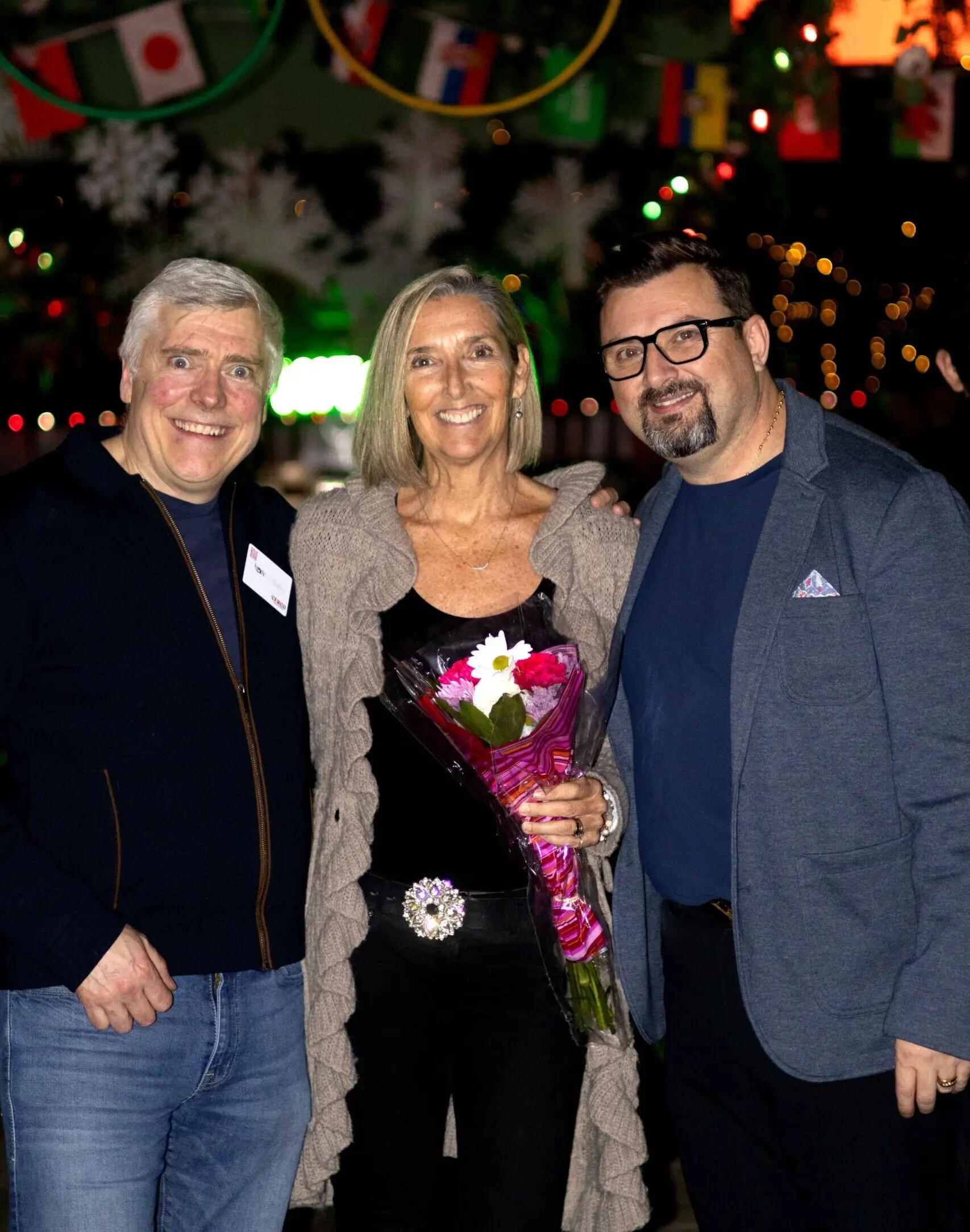 Three smiling people pose for a photo, one holding a bouquet of flowers
