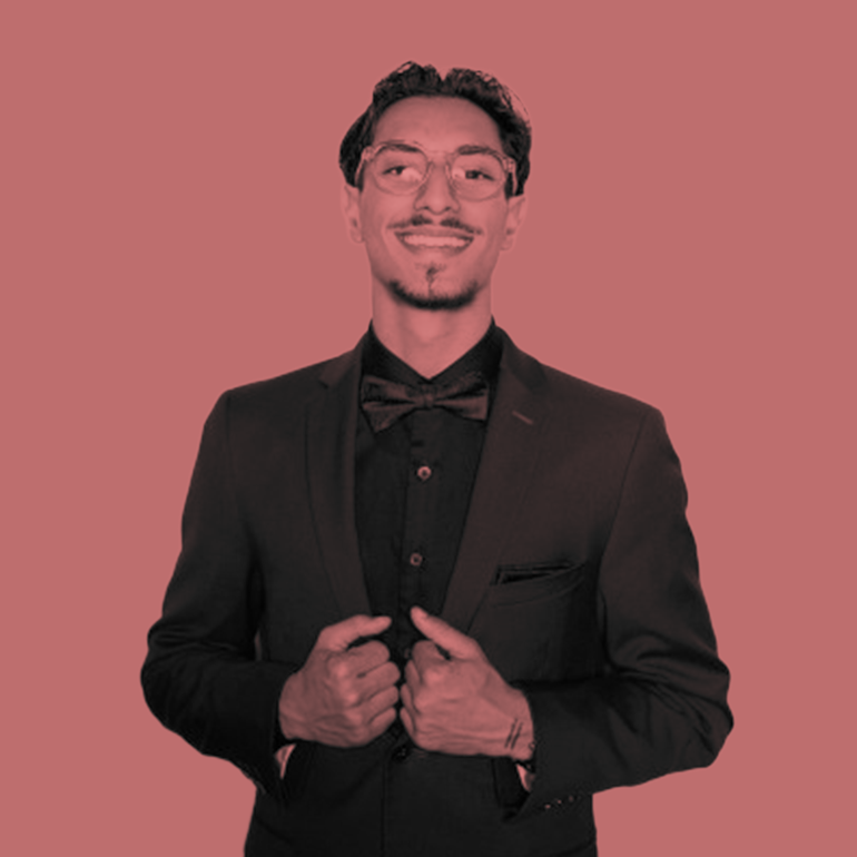 Young man in a black suit and bow tie smiles, touching his lapels against a pink background.